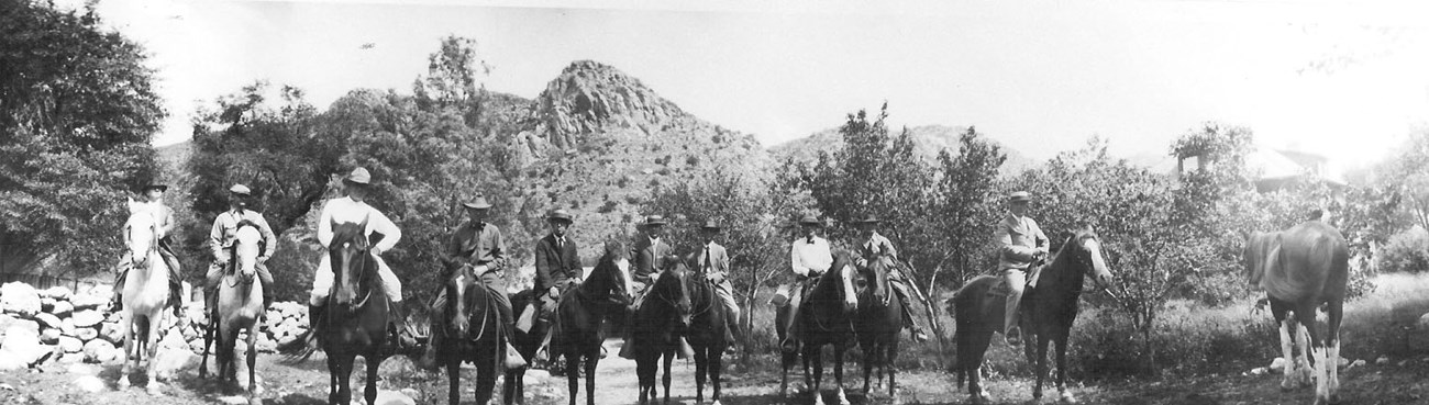 Black and white photo of ten men on horseback, with one horse on the far right, without a rider, facing away from the camera. Rocky hillside in the background.