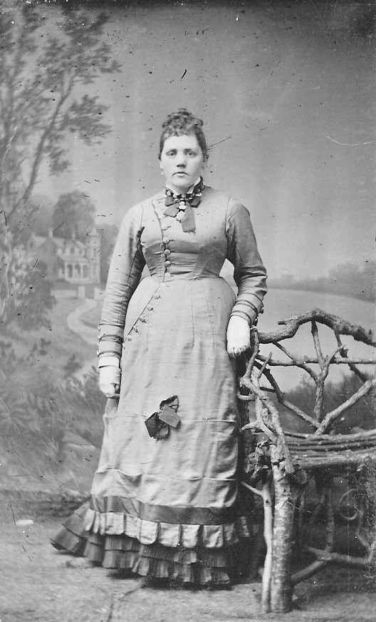 Black and white portrait of a young woman in a dress standing by a bench.