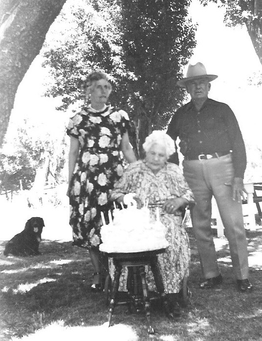 black and white photo of older woman in wheelchair, a man and a woman standing, and a large birthday cake in front.