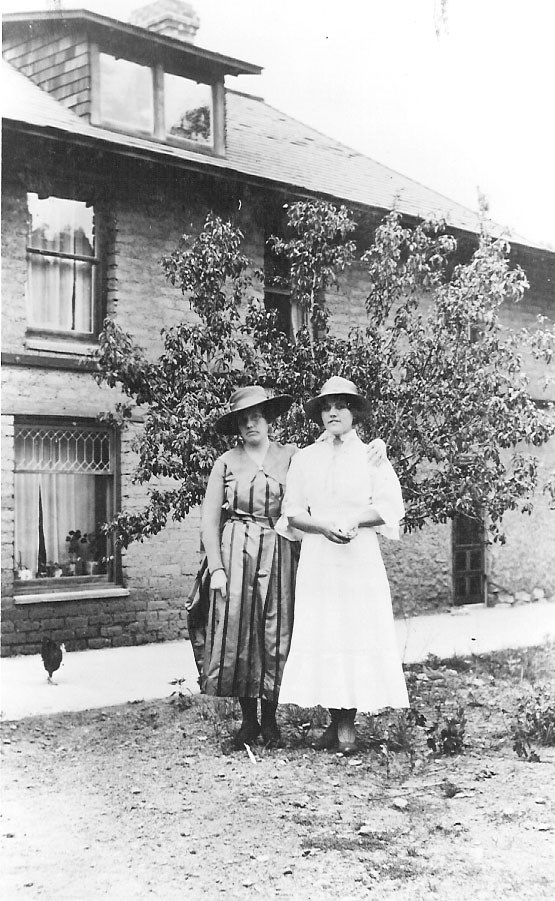 Two women wearing dresses standing in front of a two story brick house.