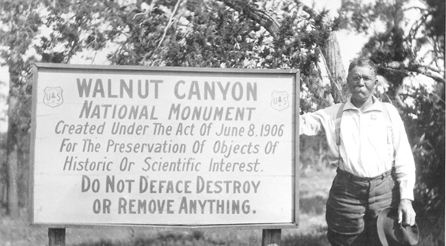 Black and white photo of a man with a mustache and hat standing by a sign about Walnut Canyon.