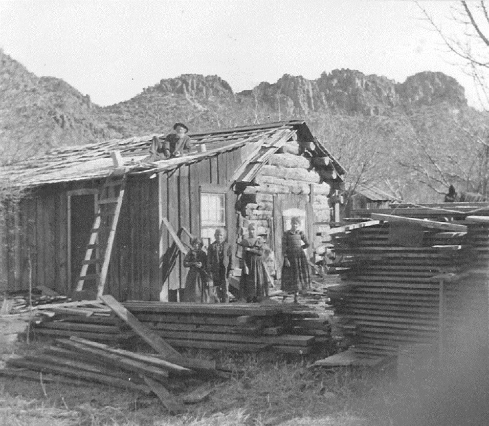 Black and white photo of man on log cabin roof, surrounded by building material and four children standing by piles of lumber.