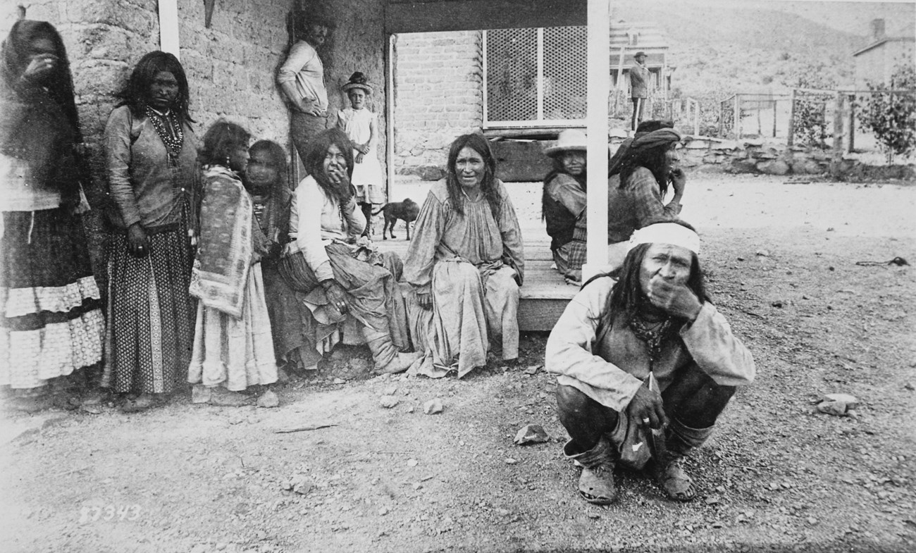 Black and white photograph of Chiricahua Apache at Fort Bowie. Many are sitting on a porch or leaning against an adobe building, and one man is squatting on the ground, wearing a headband with his hand over his mouth.