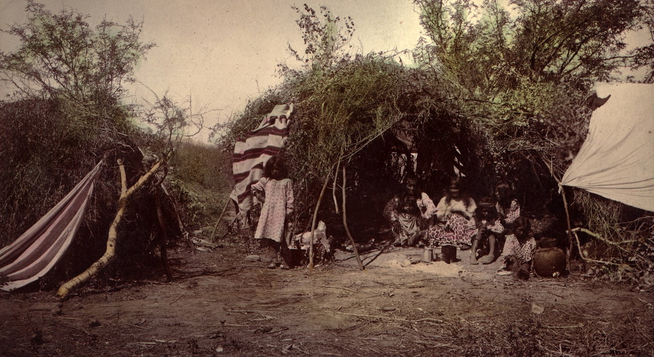 Hand-tinted photograph of wikiup, with people standing nearby, or inside.