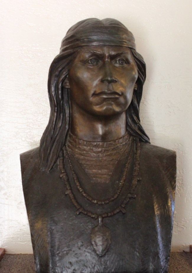 A statue bust of a man wearing a headband over his long hair, and two necklaces.