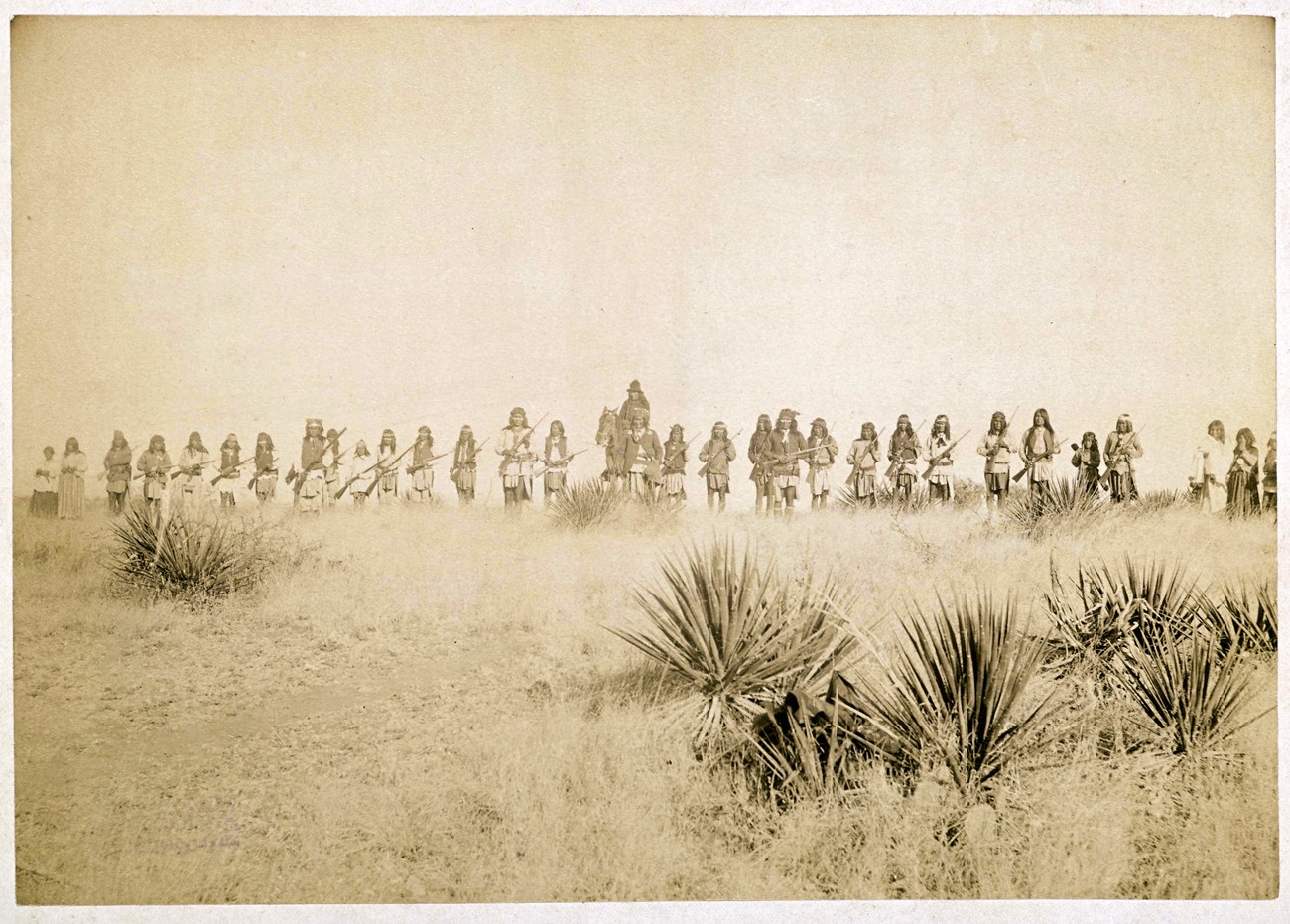 Sepia-tone photograph of line of warriors on foot, with one person on horseback. Agaves or yuccas and grasses in the foreground, and a clear sky in the background.