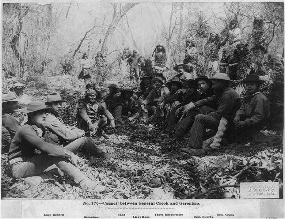 Gathering of men, mostly sitting on the ground, in a desert forest.