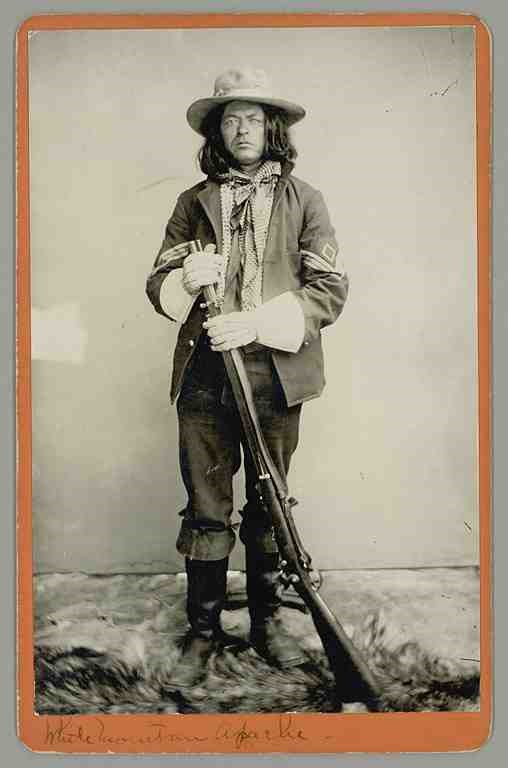 Black and white full-length portrait of young man, standing, holding a rifle, wearing a hat and military jacket over a patterned shirt.