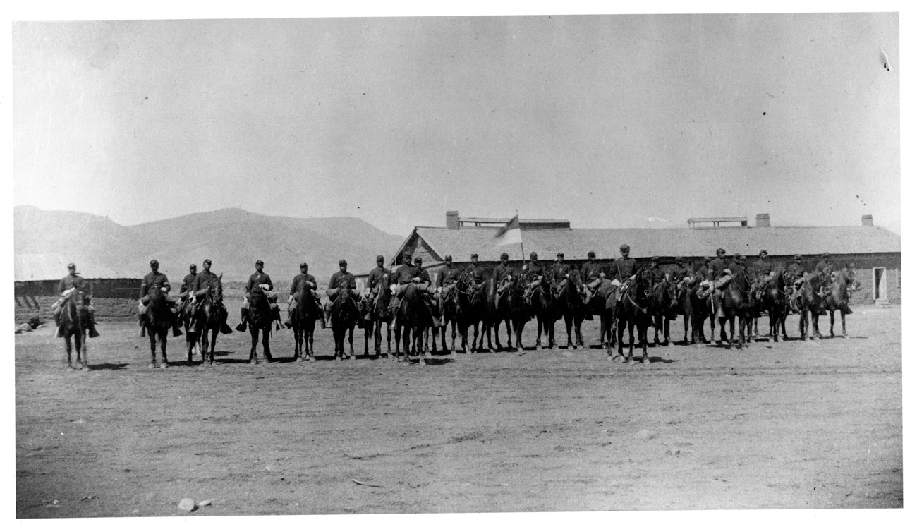 Black and white photo of African American soldiers on horses in front of building.