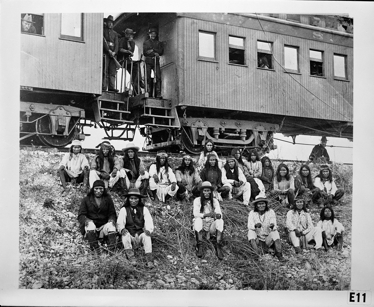 Group of men seated in front of train cars, guarded by Army soldiers standing on the train.