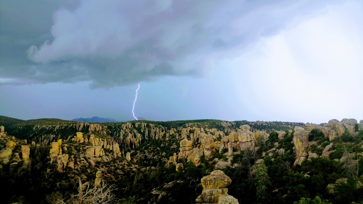 White lightning bolt striking a hill, with dark gray skies around it, and rock pinnacles in the foreground.