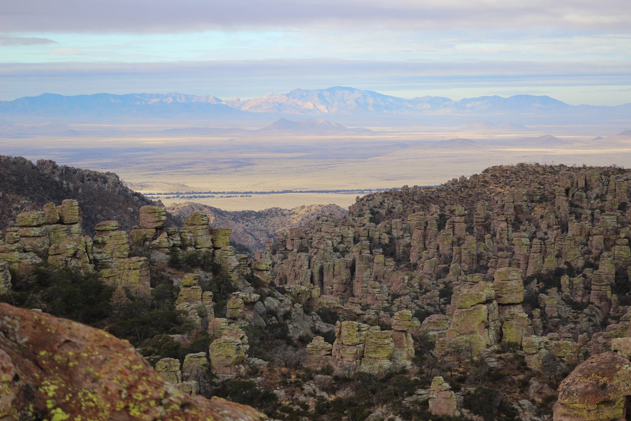 View across a valley with distant mountains on the far side. In the foreground are rock pinnacles and spires.