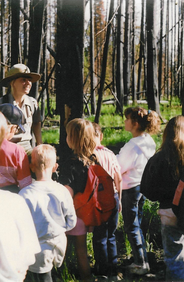 lt= A park ranger stands in front of a standing group of children in a stand of charred trees.