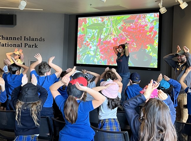 Three volunteer rangers stand at the front of the room with their hands mimicking animal ears on top of their heads as a group of students copies them.