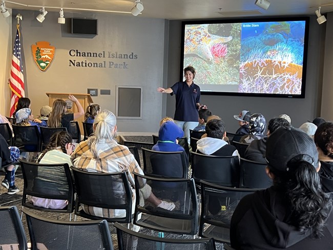 a young woman speaks in front of a group of students with images of a sea star in the background on a screen