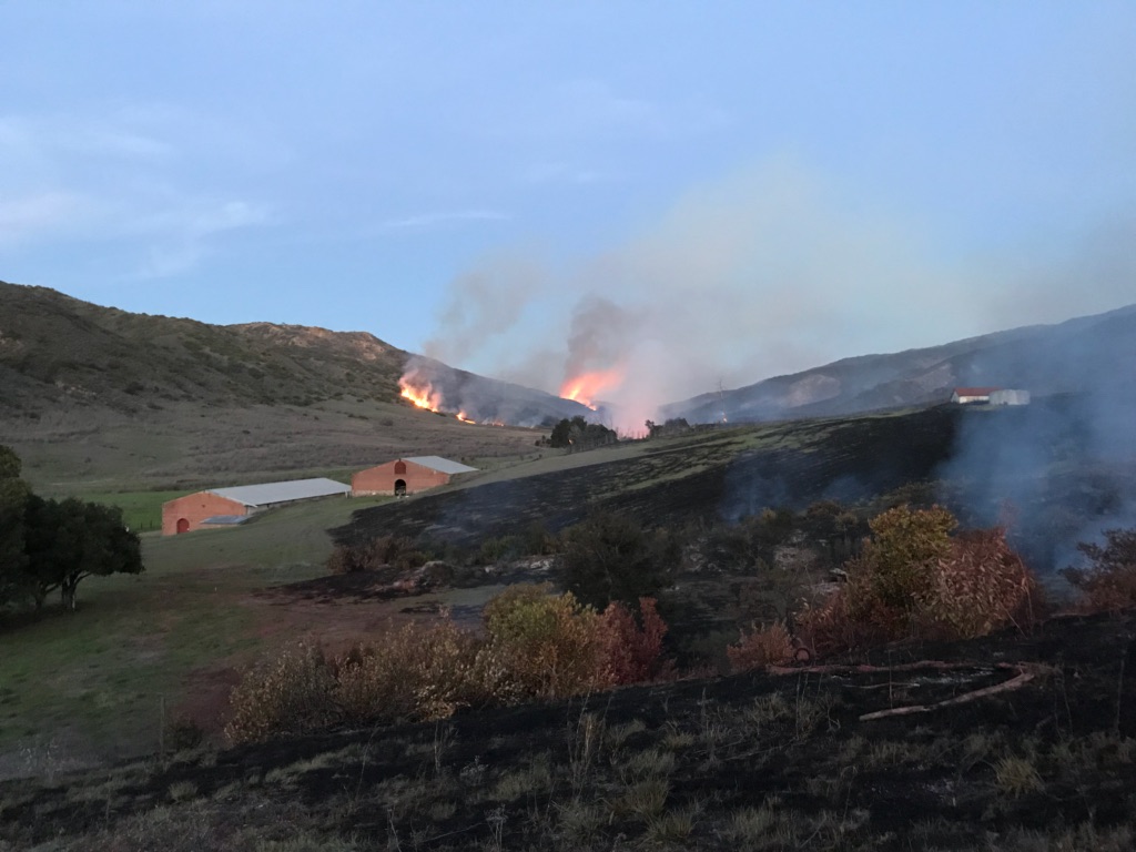 A wildifre burns through brush in the background. Structures are present in the foreground.