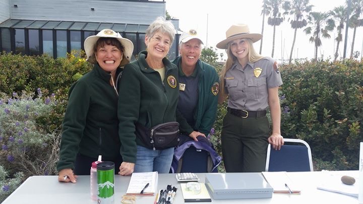 Three women in NPS volunteer uniforms pose with a female ranger at an information table. They are standing in front of a lush garden with a grey building and palm trees in the background.