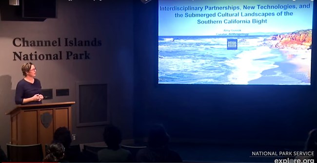 Woman at podium in theater giving lecture wtih image of coastline on screen.