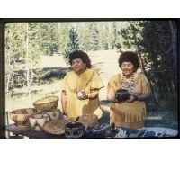 Two Native American women stand at a table holding woven baskets in front of a meadow.