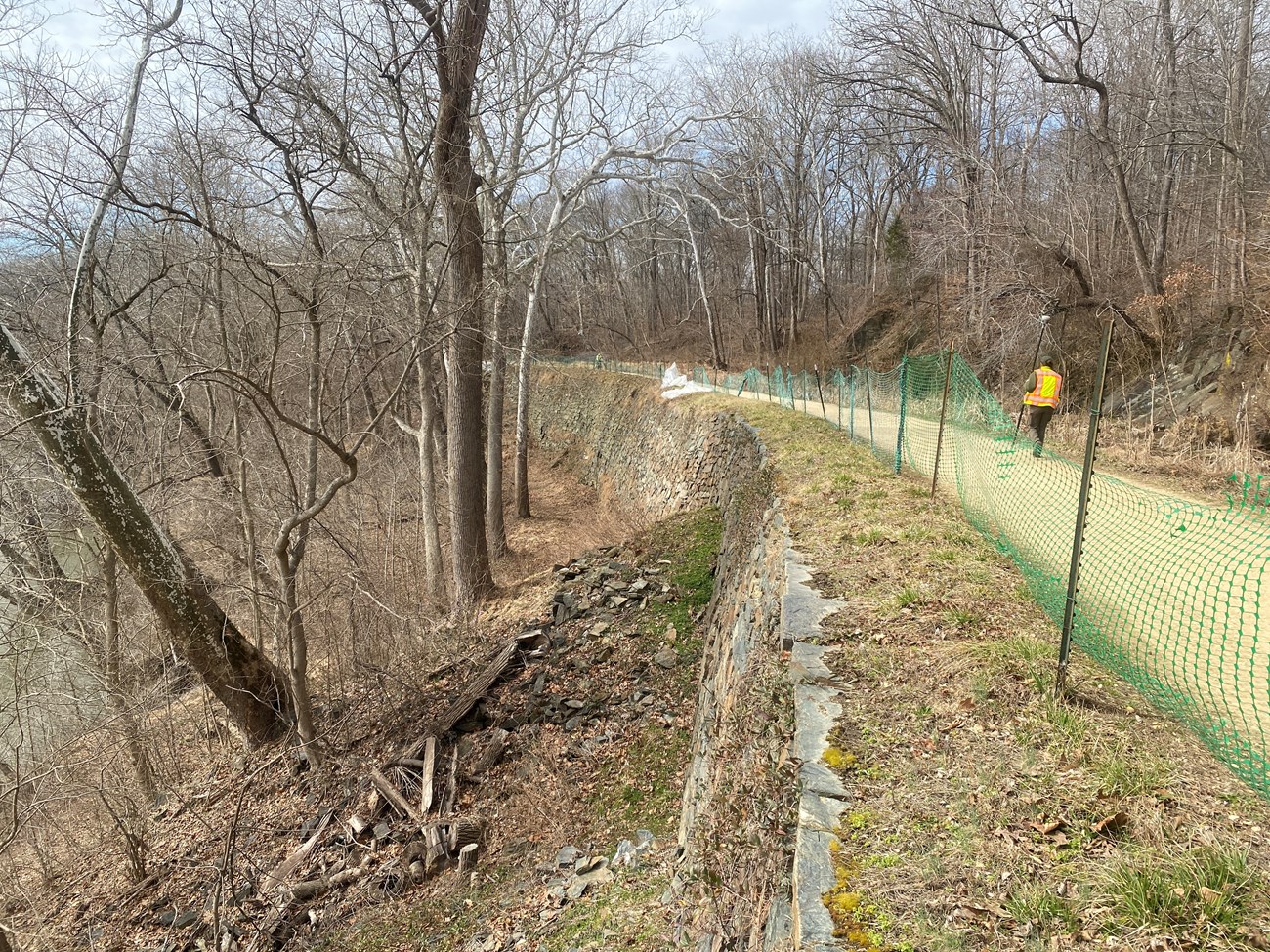 A dry-laid stone wall with safety fencing at the top next to a dirt path