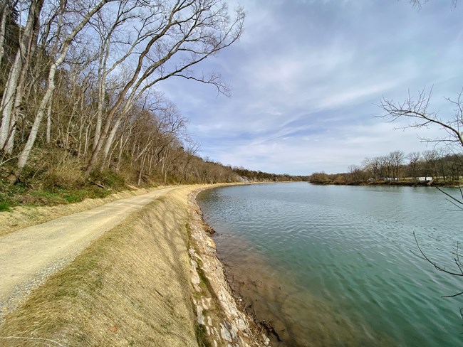 The towpath runs along a wide stretch of river with a blue sky above and trees on the left.