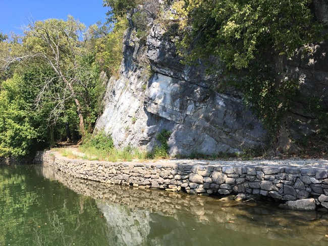 A stone retaining wall holding up a dirt pathway, winding between a river and a rock cliff