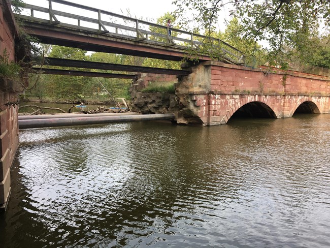 A red stone aqueduct spanning a creek with one failed arch and a bridge replacing it.