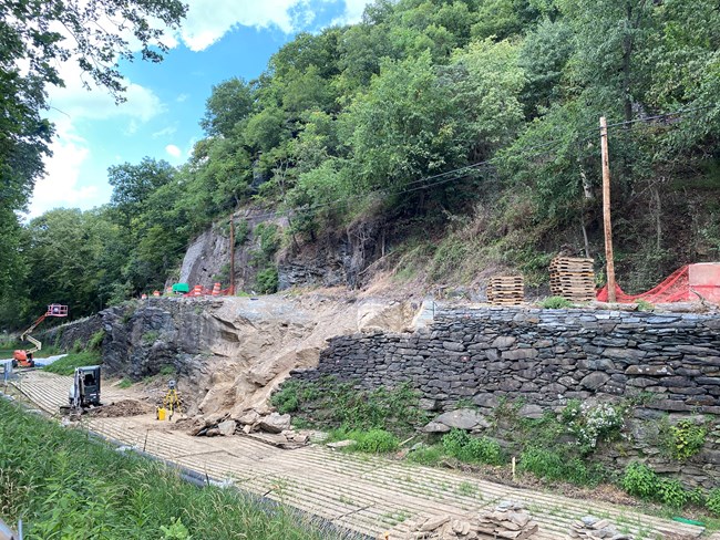 A dry-laid stone wall holding up a road has a portion of stone removed from it as part of ongoing repairs.