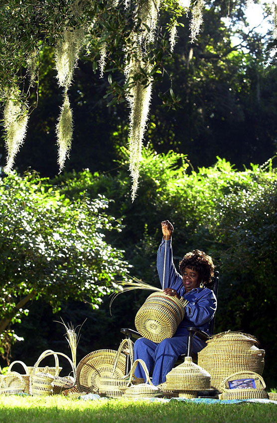 An African-American woman sews sweetgrass baskets beneath live oak trees.