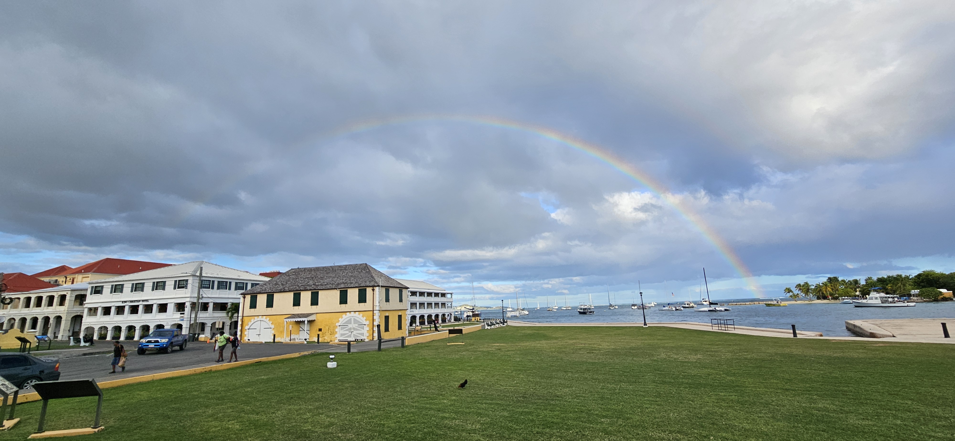 A rainbow stretches over a body of water, ships at anchor, and historic, colorful buildings.