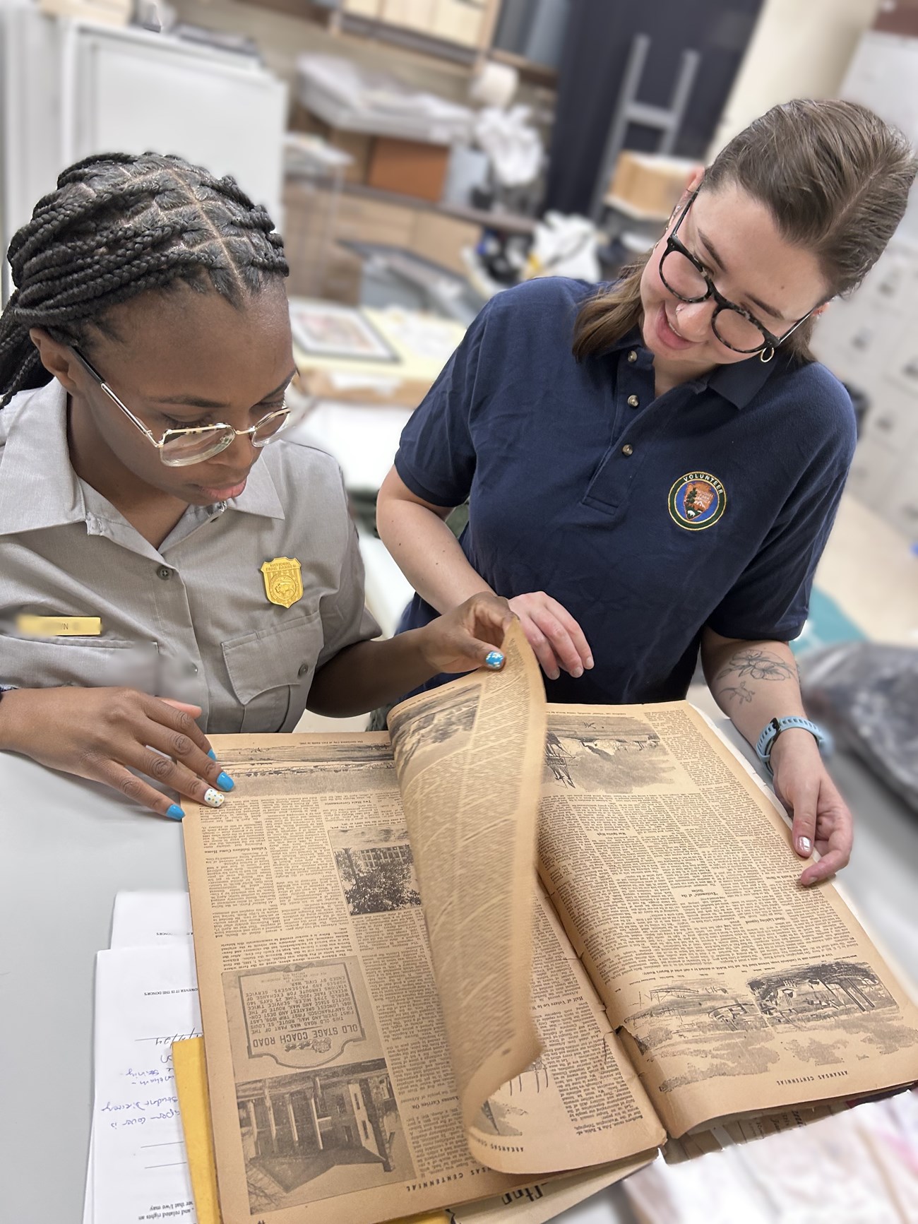 An NPS volunteer and an NPS employee review a historic newspaper together in a collections workspace, carefully turning pages and examining archival materials.