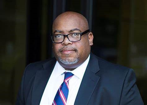 Dr. Brian Mitchell Portrait of Dr. Brian Mitchell, wearing a dark suit, white shirt, and a red and blue striped tie. He has glasses, a short beard, and a serious expression, standing in front of a blurred background.