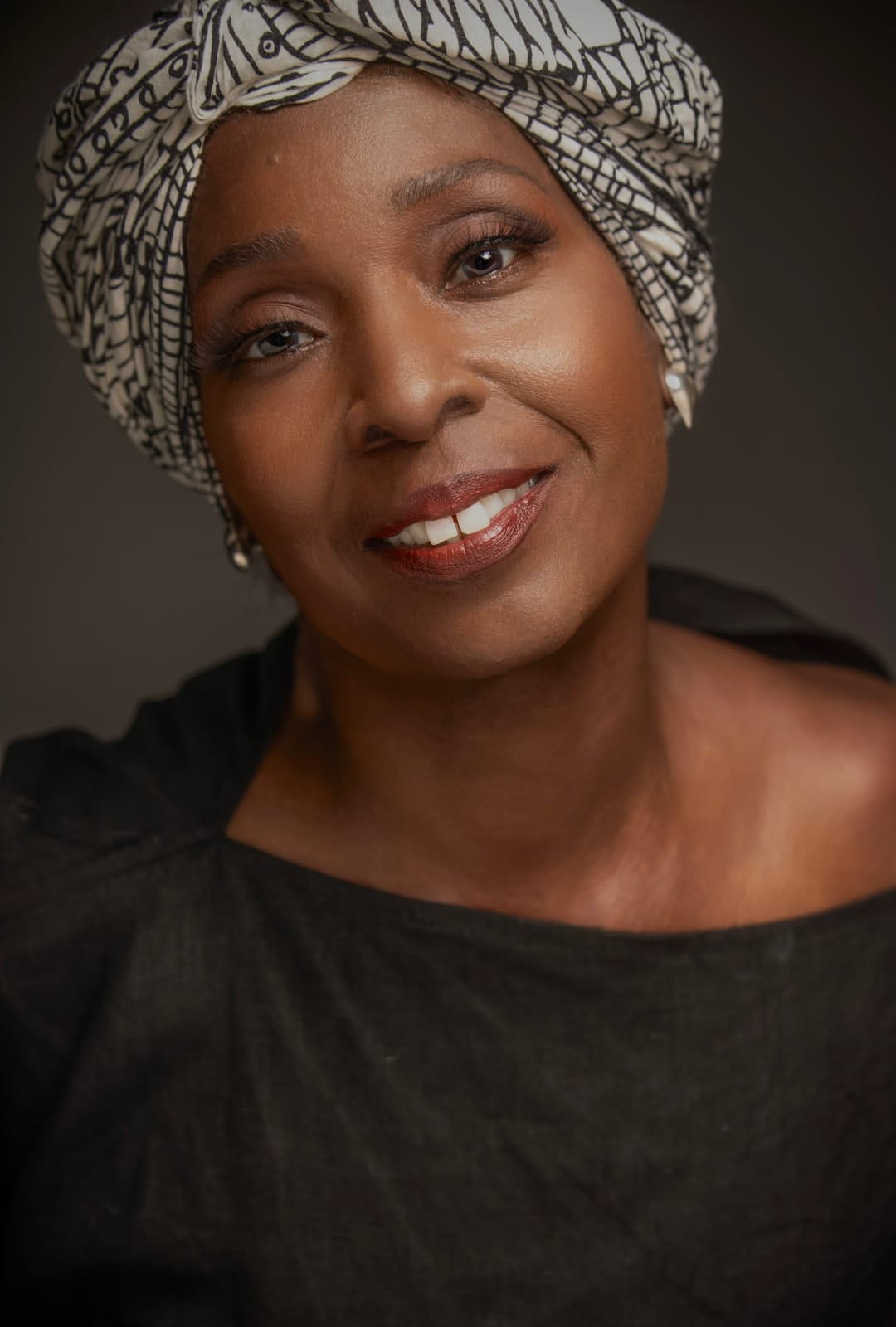 Portrait of a smiling Black woman wearing a black top and a patterned black-and-white headwrap against a dark neutral background.