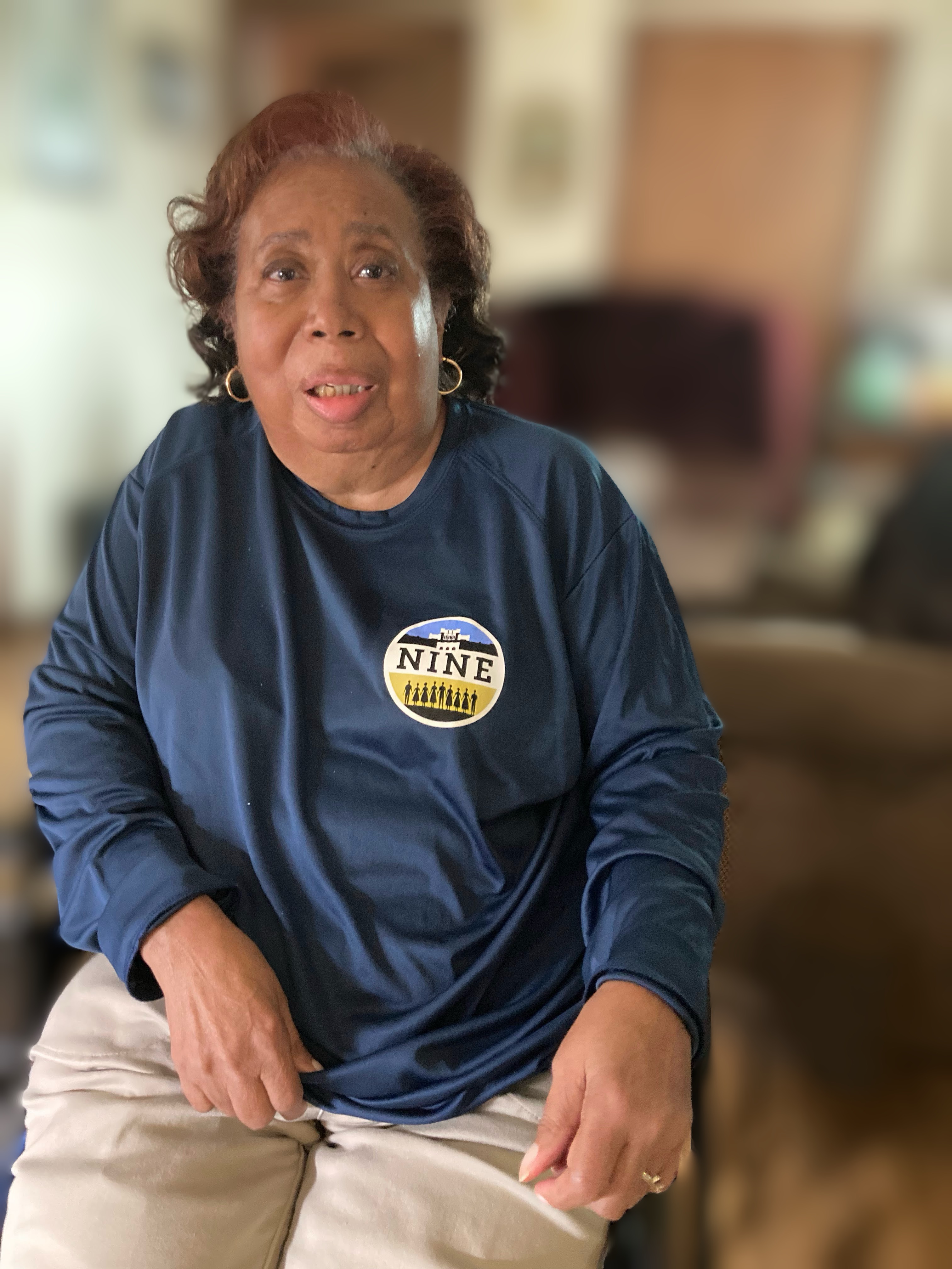 A photo of Thelma Mothershed Wair, a member of the Little Rock Nine, sitting indoors. She is wearing a navy blue shirt with a circular logo featuring the words “The Nine” and the silhouette of a group of people.