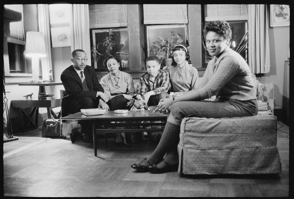 A photograph shows an African American family sitting on a sofa in their living room; an African American female teenager sits on chair adjacent to the family.