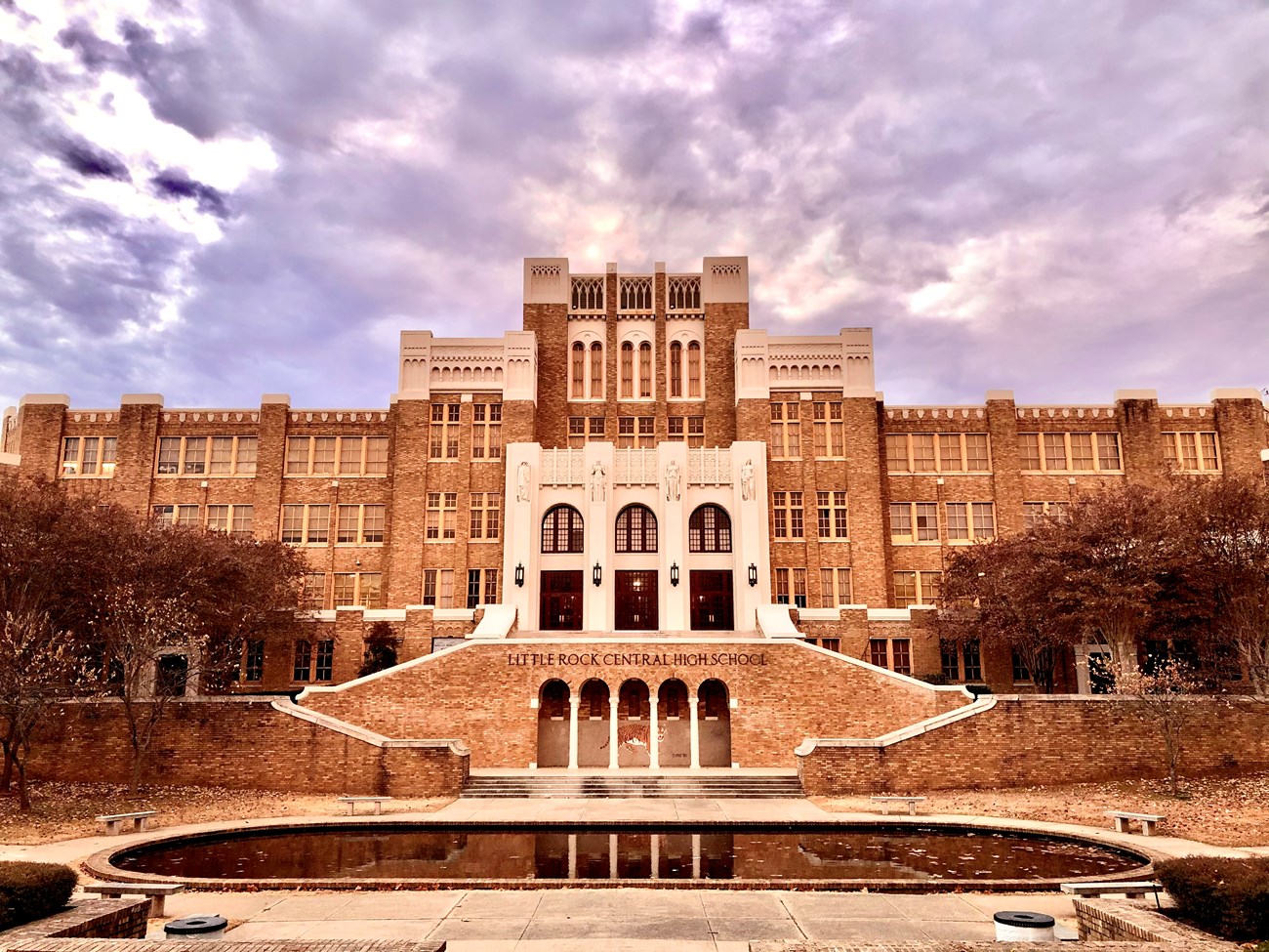 Front view of Little Rock Central High School, a large historic brick building with gothic architectural elements, under a dramatic cloudy sky. A reflecting pool and landscaped steps lead to the main entrance.