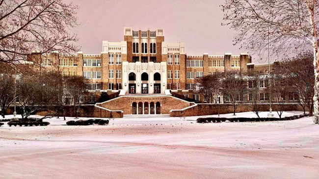 Front view of Little Rock Central High School during winter, with snow covering the ground and bare trees framing the historic brick and stone building under a soft, overcast sky.