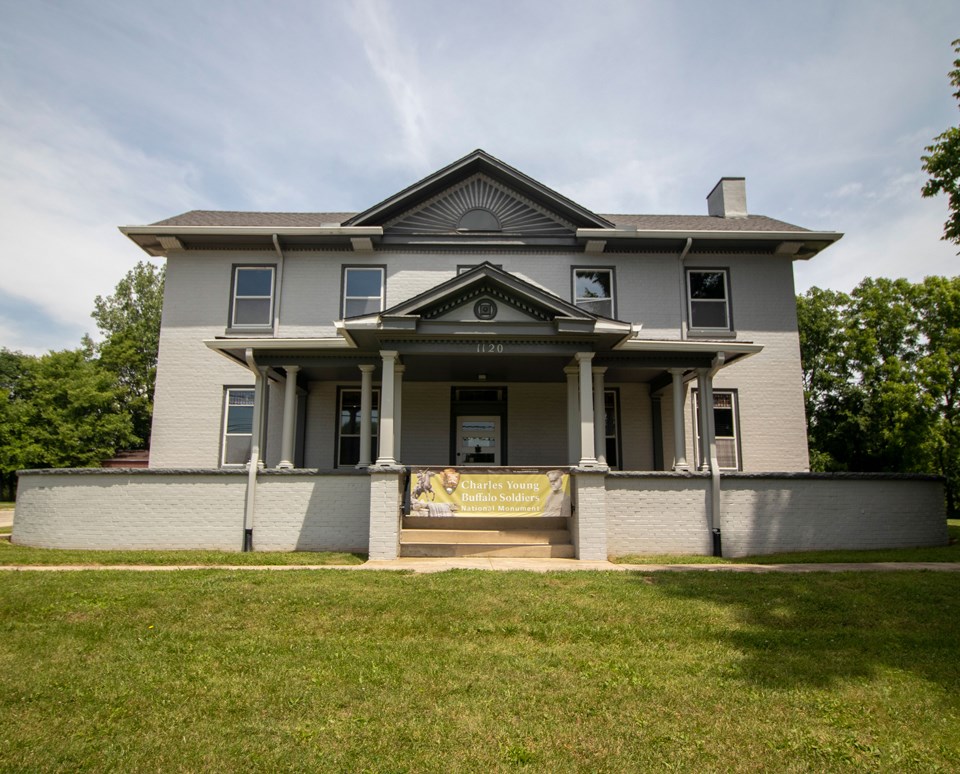 A large gray two-story house with a wide front porch on the lower level and five windows spaced equally apart on the second level and a peaked roof in the middle of the roof