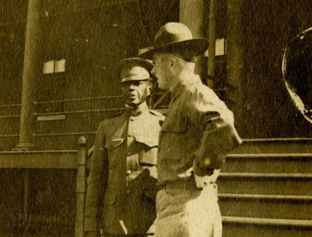 Charles Young at Camp Grant in 1919. Courtesy of the National Afro-American Museum and Cultural Center, Wilberforce, Ohio. Black and white photo of African American Army officer walking downstairs passing a white Officer. Both men are wearing World War One style uniforms and hats.