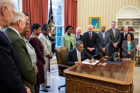 Several people stand around the President as he signs a document on his desk at the White House.