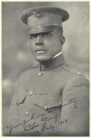 Portrait of an African American soldier wearing a short-billed cap and a dark-colored tunic. Written text reads "Yours for Race and Country, Chas. Young, 22 Feby, 1919."