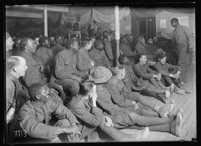 Room of Black and white male soldiers seated in rows facing toward one Black soldier who is standing up speaking