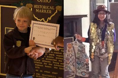 A little boy holds a paper certificate on left and a girl holds a vest full of badges next to her while wearing additional badges and a flat hat.