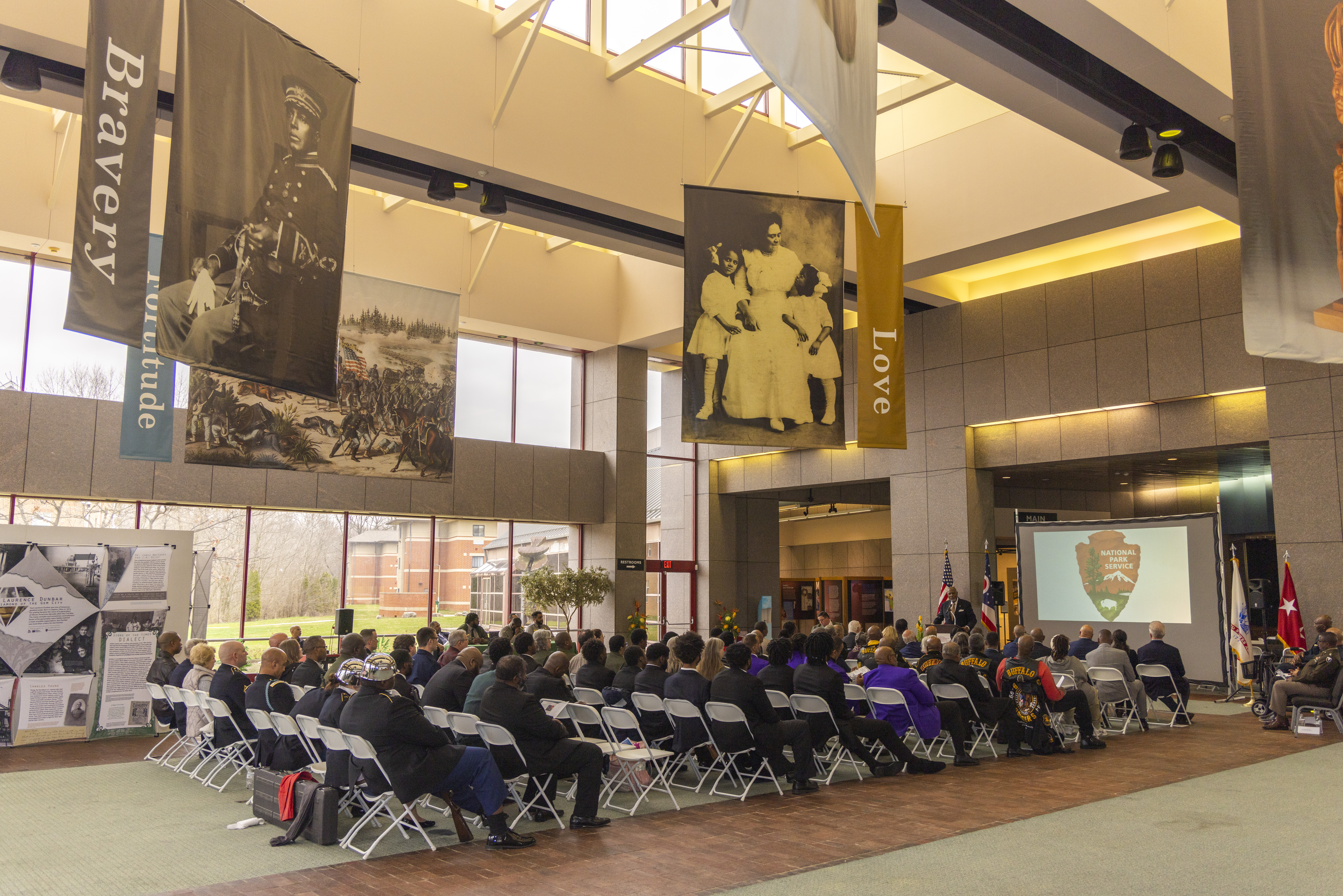 160th Birthday Event at NAAMCC Several people seated in chairs in a large hall underneath large banners susptended from the ceiling showing prominent African Americans