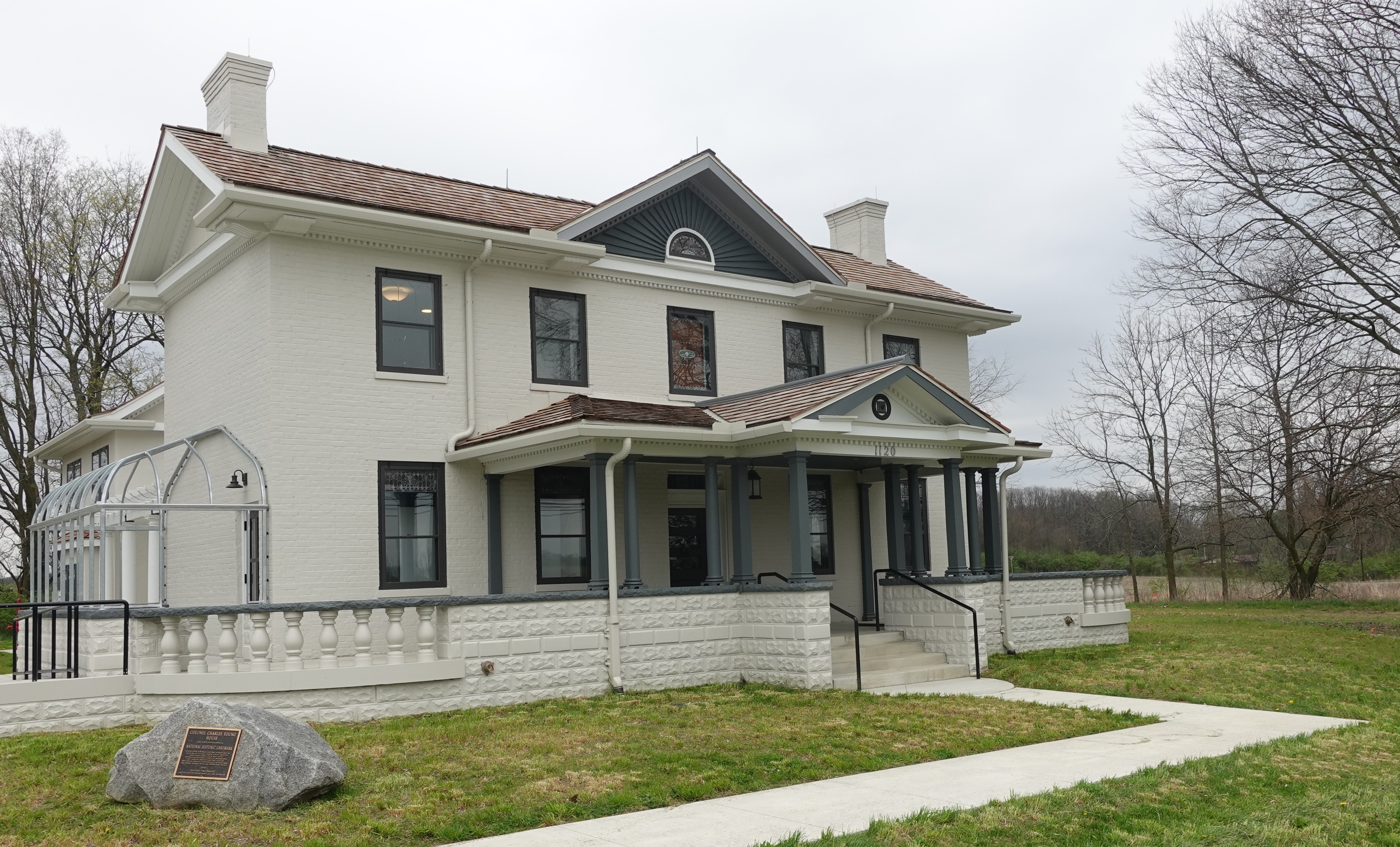 color photo of two-story brick historic home. Home is painted light gray. The front porch is covered and run the length of the front of the house. The sky in the background is cloudy.