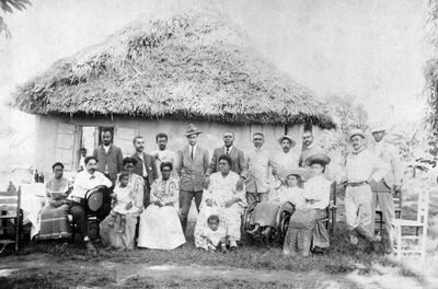 Several people standing and sitting in front of a primitive-looking hut with straw roof.