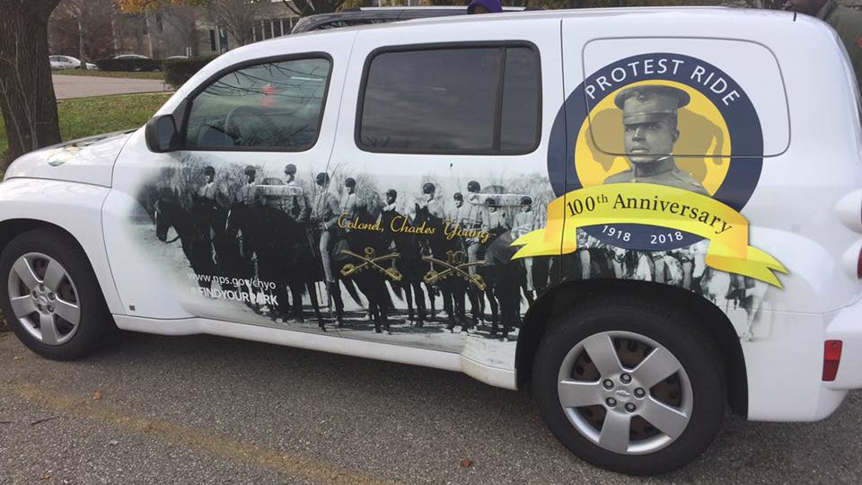 A white car with photos wrapped onto the outside showing men on horses and a yellow circle with portrait of soldier in middle with text reading PROTEST RIDE