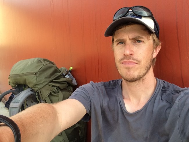 Selfie of a Caucasian man with a light beard sitting next to a backpacking bag.