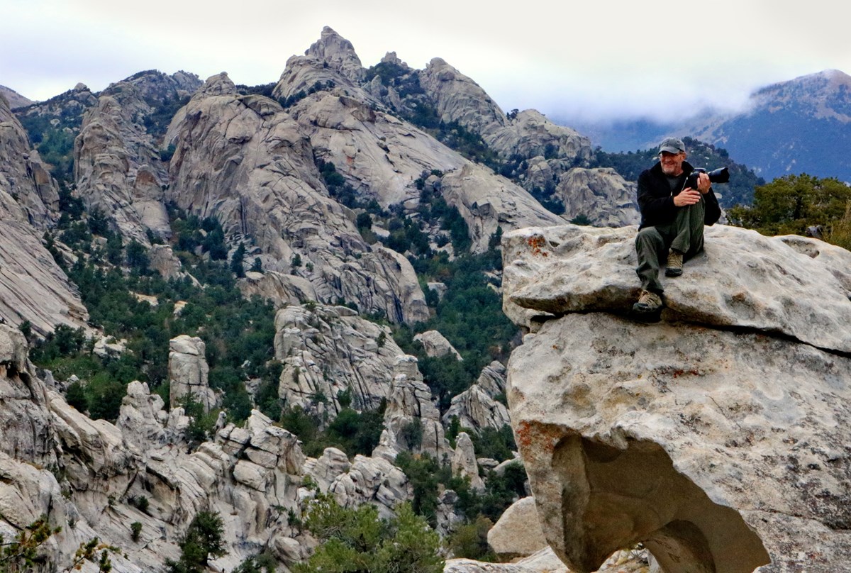 A man sits on a rock holding his camera with a grand scenic view of City of Rocks.