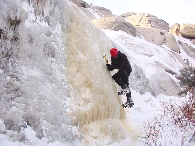 A climber ascends and ice covered rock face using crampons and an ice ax.